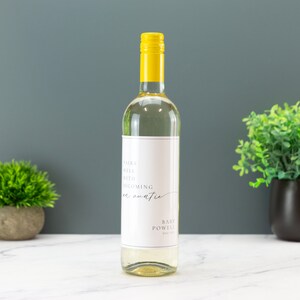 May include: A white wine bottle with a custom label that says "Pairs well with becoming an auntie" and "Baby Powell" in a minimalist font. The bottle has a yellow cap and is sitting on a white surface.
