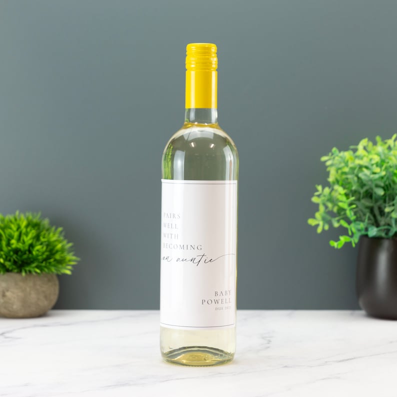 May include: A white wine bottle with a custom label that says "Pairs well with becoming an auntie" and "Baby Powell" in a minimalist font. The bottle has a yellow cap and is sitting on a white surface.