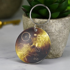 May include: A round keychain with a silver ring. The keychain features a gold and brown design with intricate line art, resembling a diagram or map. The design includes circles, lines, and geometric shapes. The keychain is likely made of metal.