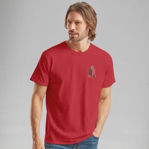 K&ouml;nnte beinhalten: Ein rotes T-Shirt mit Rundhalsausschnitt und einer kleinen Grafik auf der linken Brust. Die Grafik zeigt ein stilisiertes Geb&auml;udedesign in Grau und Rot mit dem Wort "Fairview" darunter. Das Shirt ist aus einem weichen Material.