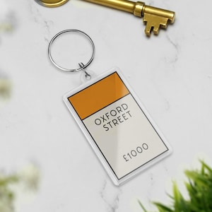 Op de afbeelding: Een heldere rechthoekige sleutelhanger met een zilveren ring, met een Monopoly Oxford Street-ontwerp. Het bovenste gedeelte is oranje, met "OXFORD STREET" en "£1000" gedrukt op de witte achtergrond. Een gouden sleutel staat op de achtergrond.