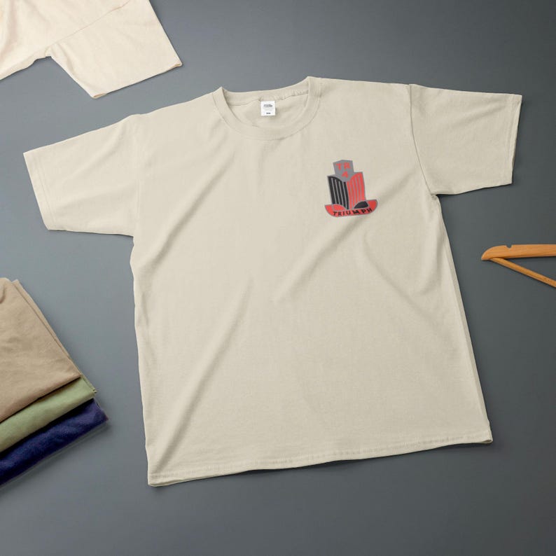 K&ouml;nnte beinhalten: Cremefarbenes T-Shirt mit einer kleinen Grafik auf der Brust. Die Grafik zeigt ein stilisiertes Design in Rot, Schwarz und Grau mit dem Wort "TRIUMPH" darunter. Zus&auml;tzliche gefaltete Hemden sind im Bild zu sehen.