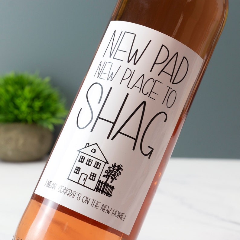 Puede incluir: Una botella de vino rosado con una etiqueta blanca que dice "NEW PAD NEW PLACE TO SHAG" con una peque&ntilde;a ilustraci&oacute;n de una casa y el texto "I MEAN CONGRAT'S ON THE NEW HOME!"
