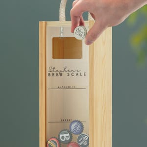 May include: Wooden box with a clear glass front, labeled "Stephen's Beer Scale" with the categories "Alcoholic" and "Expert". The box is filled with bottle caps.