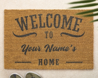 Puede incluir: Felpudo rectangular marrón con las palabras "WELCOME TO Your Name's HOME" en letras negras. La alfombra está hecha de fibra natural. Un par de zapatillas de camuflaje se ven en la parte inferior.