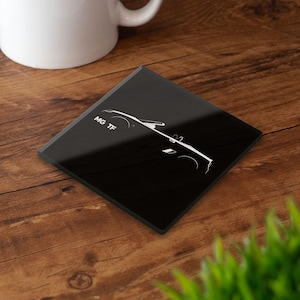 Puede incluir: Un posavasos cuadrado negro con un contorno blanco de un coche y el texto "MG TF". El posavasos está sobre una superficie de madera, con una taza blanca en el fondo.