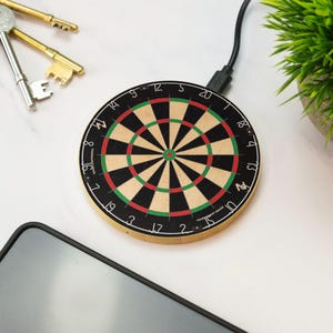 Könnte beinhalten: Ein runder kabelloser Ladegerät mit Dartscheiben-Design. Das Ladegerät hat ein schwarzes, grünes und rotes Zielmuster mit Zahlen am Rand. Ein schwarzes Ladekabel ist angeschlossen. Ein Smartphone und Schlüssel sind ebenfalls sichtbar.