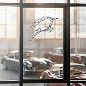 May include: A chrome-colored leaping deer emblem, likely a car badge, is affixed to a window. Classic cars are visible in the background, suggesting a vintage or luxury automotive theme. The emblem's design is sleek and modern.