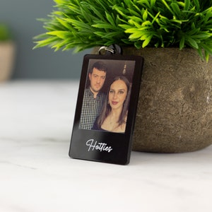 May include: Black acrylic keychain with a photo of a couple and the text "Hotties" printed in white.