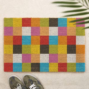May include: A rectangular doormat with a colorful checkerboard pattern. The squares are various colors, including red, yellow, blue, green, and pink. The mat is made of a textured material, and a pair of camouflage sneakers are at the bottom.