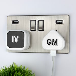 Op de afbeelding: Een wit stopcontact met twee USB-poorten en twee standaard stopcontacten. Een witte USB-adapter met de tekst "IV" is aangesloten op een van de stopcontacten. Een witte USB-adapter met de tekst "GM" is aangesloten op het andere stopcontact.