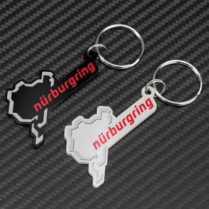 May include: Two keyrings with a racetrack outline design. The word "nürburgring" is in red. One keyring is black with a white outline, the other is white with a black outline. Both have silver key rings.