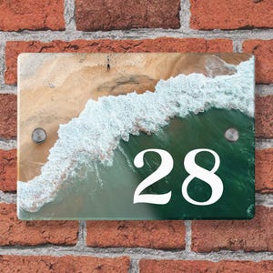 Peut inclure: Plaque de numéro de maison rectangulaire avec une scène de plage, des vagues blanches et le numéro 28 en blanc. La plaque est fixée sur un mur de briques rouges avec deux vis argentées.