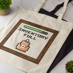 Op de afbeelding: Een beige canvas tas met een pixel art afbeelding van een bruine rugzak in een bruin kader. Boven de rugzak staat de tekst INVENTORY FULL in zwarte pixelletters, vergezeld van een groene laadbalk aan de bovenzijde van het kader.