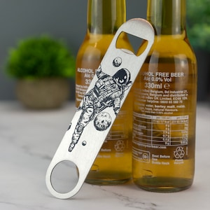 May include: A silver bottle opener with a black and white illustration of an astronaut floating in space. The astronaut is wearing a spacesuit and helmet. The bottle opener has a hole in the top for hanging.