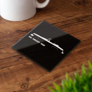 Puede incluir: Posavasos cuadrado negro con un dibujo lineal blanco de un coche descapotable. El texto "MG MIDGET 1500" está impreso en blanco en el lado izquierdo. El posavasos está sobre una superficie de madera.