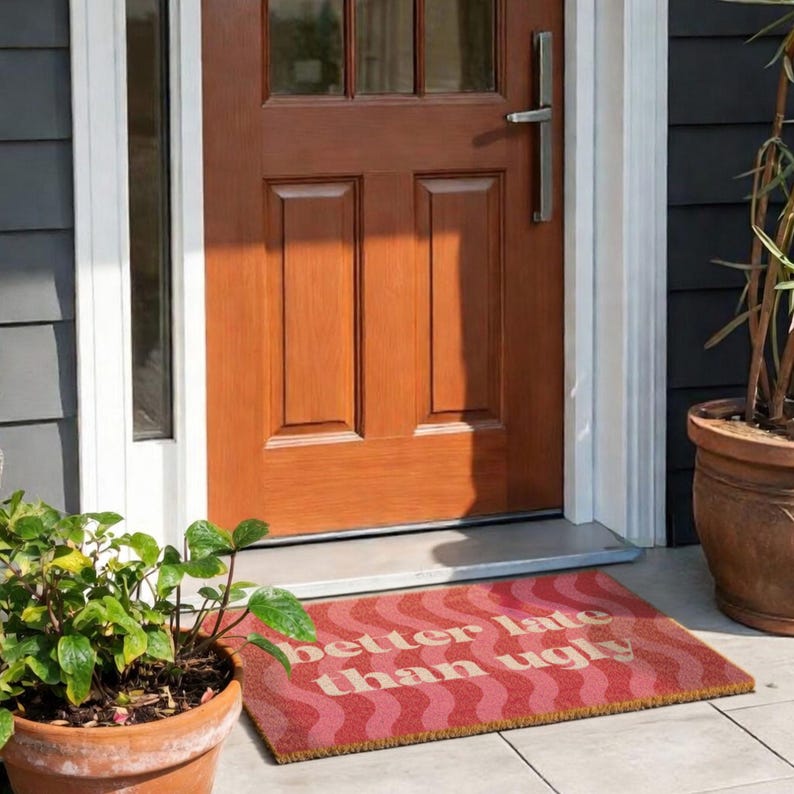 May include: A rectangular doormat with a red and pink wavy pattern and the text "better late than ugly" in a light pink color. The mat is placed in front of a brown wooden door.