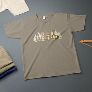 Könnte beinhalten: Ein taupefarbenes T-Shirt mit kurzen Ärmeln und Rundhalsausschnitt, das einen Grafikdruck von weißen Kiefern und den braunen Schriftzug I GOT WOOD in einer blockartigen, texturierten Schrift in der Brustmitte aufweist. Standardpassform mit Rippkragen.