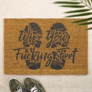Könnte beinhalten: Eine rechteckige Fußmatte aus natürlicher brauner Kokosfaser. Die Matte zeigt eine schwarze Grafik von zwei Fußabdrücken und den Text "Wipe Your Fucking Feet" in einer stilisierten Schriftart. Ein Paar Camouflage-Sneaker sind unten.