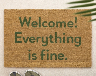 Op de afbeelding: Een rechthoekige beige deurmat van kokosvezel met donkergroene tekst die luidt Welcome! Everything is fine. De mat heeft een grove, vezelige textuur en ligt op een lichte ondergrond naast een paar sneakers met een camouflagepatroon.