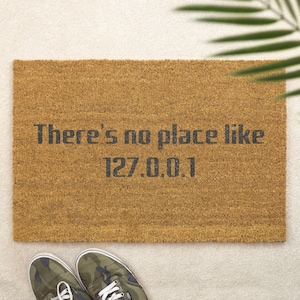 May include: A rectangular doormat made of natural brown coir fiber. The mat features the phrase "There's no place like 127.0.0.1" in dark gray block letters. A pair of camouflage sneakers is visible at the bottom.