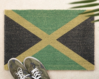 Pode incluir: Um tapete retangular com o design da bandeira jamaicana em preto, verde e dourado. Um par de tênis camuflados com cadarços brancos são colocados em frente ao tapete. O tapete é feito de um material texturizado.