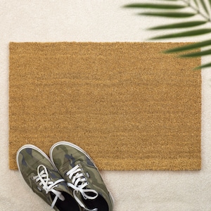 May include: A rectangular brown coir doormat with a textured surface. A pair of green camouflage canvas sneakers with white laces are placed in front of the doormat. The scene is set on a light-colored rug.