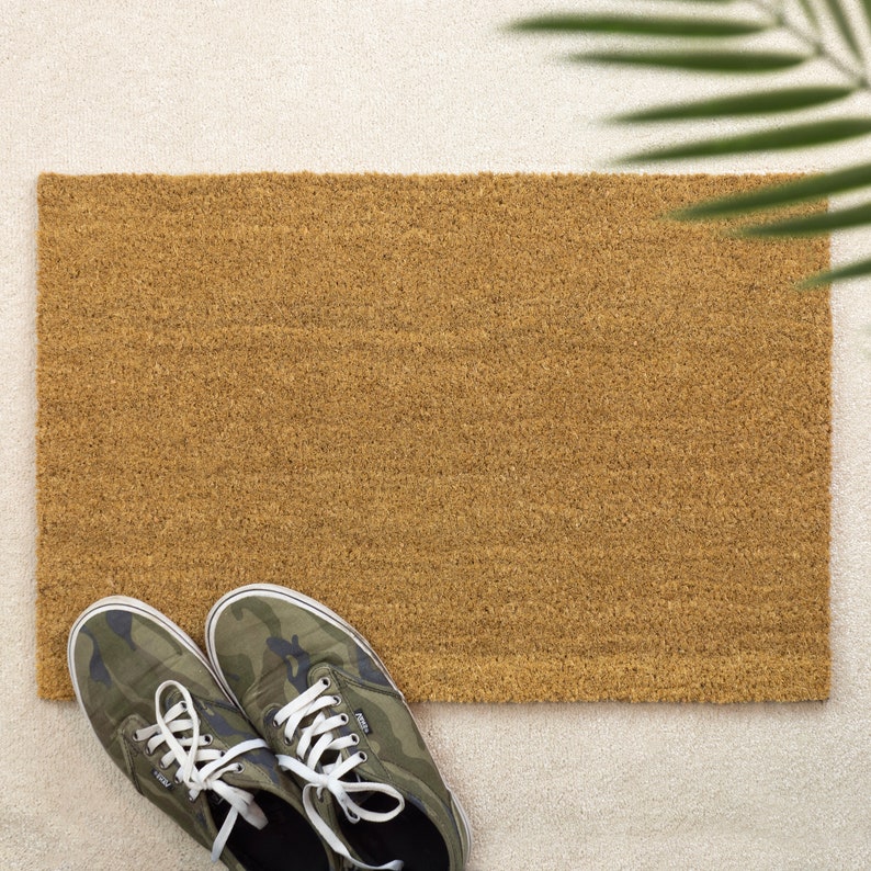 May include: A rectangular brown coir doormat with a textured surface. A pair of green camouflage canvas sneakers with white laces are placed in front of the doormat. The scene is set on a light-colored rug.