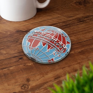 May include: A round coaster with a globe design. The coaster features a blue background with red continents and a silver outline. A white mug is in the background, and the coaster is on a wooden surface.