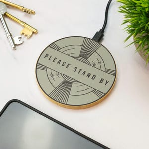 以下が含まれることがあります： セージグリーンの円形ワイヤレス充電パッド。黒い電源コードが付いています。表面には、ヴィンテージのテレビのテストパターンを模したグラフィックデザインが施されており、中央に黒い文字で「PLEASE STAND BY」と印刷されています。