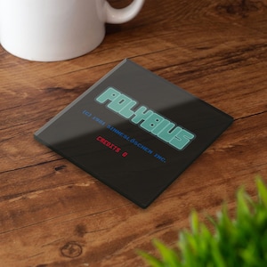 Op de afbeelding: Vierkante zwarte onderzetter met het woord "POLYBIUS" in teal, met de tekst "(c) 1981 SINNESLÖSCHEN INC." en "CREDITS 0" in rood. De onderzetter ligt op een houten oppervlak.