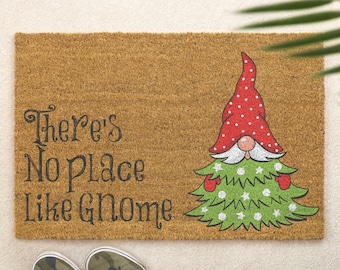 Può includere: Un zerbino rettangolare con la scritta "There's No Place Like Gnome" e un design a forma di gnomo albero di Natale. Lo gnomo ha un cappello rosso a pois bianchi, un naso rosa e guanti rossi. Lo zerbino è marrone e ha una superficie strutturata.