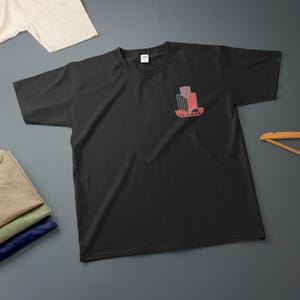 K&ouml;nnte beinhalten: Schwarzes T-Shirt mit Rundhalsausschnitt und einem roten und grauen Aufdruck auf der linken Brust. Die Grafik zeigt ein stilisiertes Design mit dem Wort "TRIUMPH" darunter. Zus&auml;tzliche gefaltete Hemden sind im Hintergrund zu sehen.