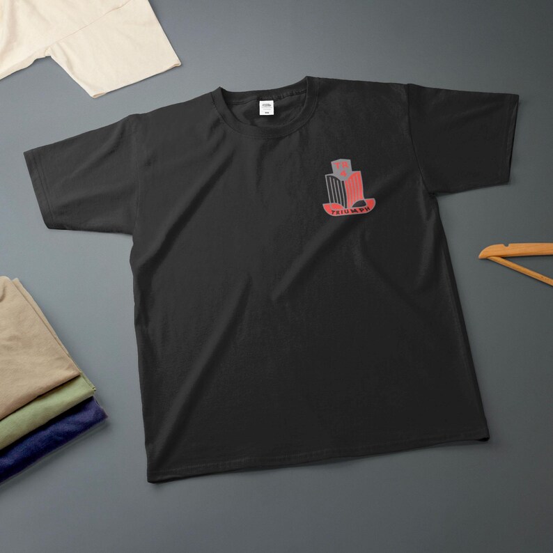 K&ouml;nnte beinhalten: Schwarzes T-Shirt mit Rundhalsausschnitt und einem roten und grauen Aufdruck auf der linken Brust. Die Grafik zeigt ein stilisiertes Design mit dem Wort "TRIUMPH" darunter. Zus&auml;tzliche gefaltete Hemden sind im Hintergrund zu sehen.
