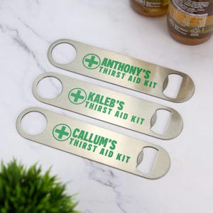 May include: Three silver bottle openers with the text "ANTHONY'S THIRST AID KIT", "KALEB'S THIRST AID KIT", and "CALLUM'S THIRST AID KIT" in green. Each opener has a circular hole and a bottle cap opener. A green cross is printed on each opener.