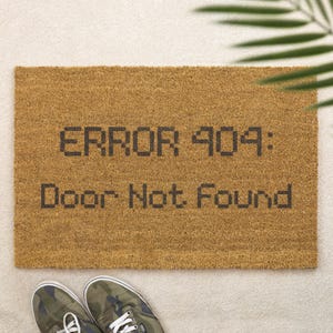 May include: A rectangular tan coir doormat featuring the black pixelated text ERROR 404: Door Not Found. The mat has a coarse, fibrous texture and is positioned above a pair of camouflage patterned trainers with white laces and rubber soles.