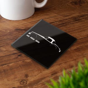 Puede incluir: Posavasos cuadrado negro con un contorno blanco de un coche MG MGA 1500. El texto "MG MGA 1500" está impreso en blanco en el posavasos. El posavasos está sobre una superficie de madera.