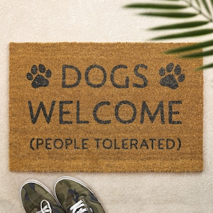 May include: A rectangular doormat made of natural brown coir fibre. The mat features the text "DOGS WELCOME (PEOPLE TOLERATED)" in black, with paw print graphics. Camouflage trainers are visible at the bottom.