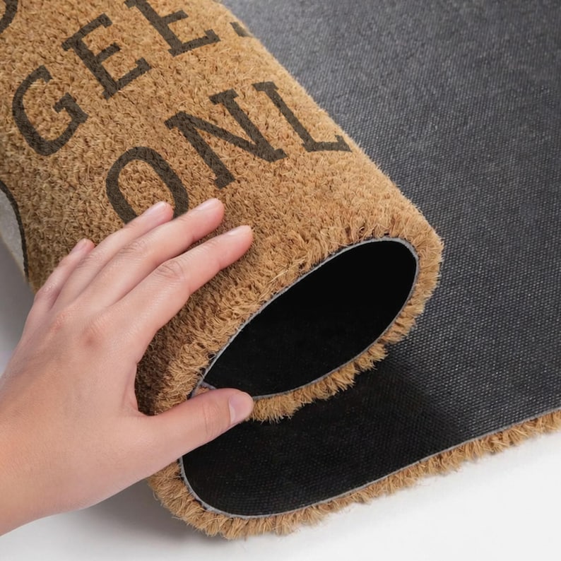 May include: A rolled-up brown coir doormat with the text "GEE ONLY" in dark brown lettering. The mat has a black backing and a fringed edge. The image shows a hand partially unrolling the mat.