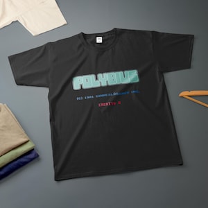 Pode incluir: Uma t-shirt preta de manga curta com o texto pixelizado azul claro POLYBIUS, o texto azul (C) 1981 SIMMESLÖSCHEN INC. e o texto vermelho CREDITS 0 impressos na frente. A t-shirt tem gola redonda e um design de corte padrão.