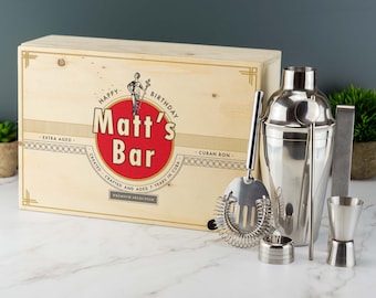 May include: A wooden box with a personalised label that says "Happy Birthday Matt's Bar" and a set of stainless steel cocktail shaker and bar tools. The box is made of wood and has a natural finish. The label is printed with black text on a white background. The cocktail shaker is made of stainless steel and has a silver finish. The bar tools include a jigger, a strainer, a muddler, and a bar spoon.
