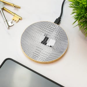 Op de afbeelding: Een zilveren draadloze oplader in de vorm van een discobal met een houten rand. De oplader is aangesloten op een zwarte USB-kabel. Een smartphone en sleutels zijn ook zichtbaar.