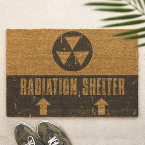 Puede incluir: Una alfombra rectangular hecha de fibras naturales. La alfombra presenta un símbolo de radiación negro y las palabras "RADIATION SHELTER" en negrita. Dos flechas apuntan hacia arriba. Zapatillas de camuflaje en la parte inferior.
