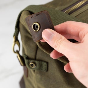 May include: Close-up of a green canvas duffel bag with a brown leather strap and a brass buckle. The strap has a brass grommet.