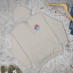 May include: A light beige hoodie with a front pocket and a hood. The hoodie features a small, colourful graphic of a globe with text. A wooden hanger and a pair of jeans are visible in the background.