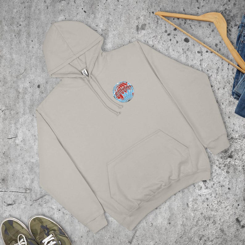 May include: A light beige hoodie with a front pocket and a hood. The hoodie features a small, colourful graphic of a globe with text. A wooden hanger and a pair of jeans are visible in the background.
