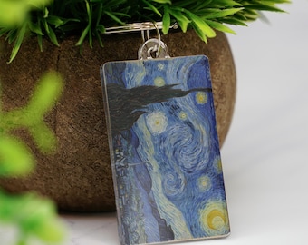 May include: A rectangular keychain featuring a print of Van Gogh's "Starry Night" painting. The keychain has a silver-colored metal ring and a clear plastic casing. The artwork depicts a swirling blue night sky with yellow stars and a dark silhouette of a town.