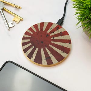 May include: A circular wireless charging pad featuring a distressed red and beige sunburst pattern. The device is connected to a black power cable and displays a weathered, vintage-style graphic design across its flat, round surface.