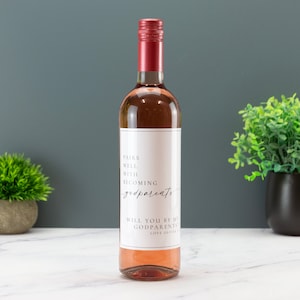 May include: A bottle of rose wine with a white label that says "Pairs well with becoming godparents. Will you be my godparents? Love Olivia."