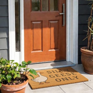 May include: A rectangular doormat with the text "SILLY GEESE ONLY" and a cartoon goose illustration. The mat is brown and sits in front of a brown wooden door. Potted plants frame the door.